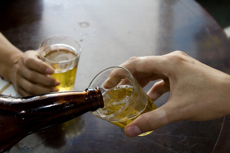 Foto de uma mão segurando um copo de cerveja. Uma garrafa está enchendo o copo