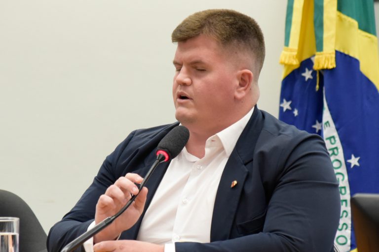 Deputado Felipe Rigoni fala ao microfone. Ele é branco, tem os olhos fechados e usa terno escuro