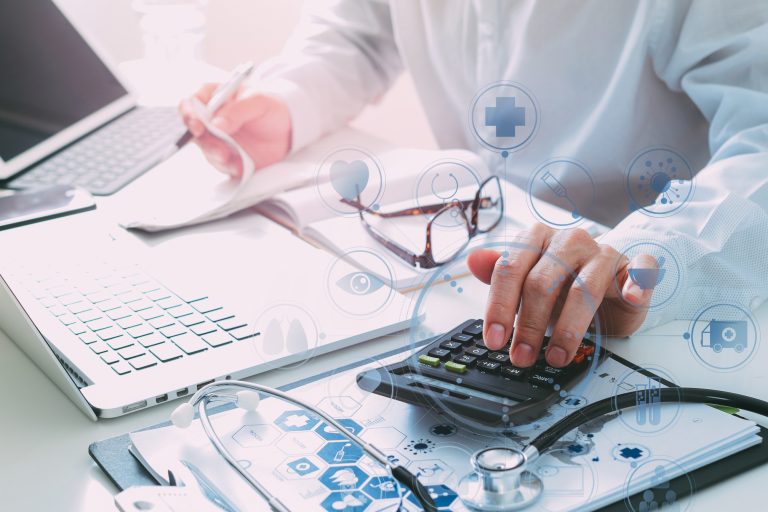 Foto de um médico mexendo no computador. Em cima da mesa estão uma calculadora, um estetoscópio e gráficos, 