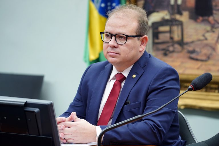 Deputado Luiz Carlos está sentado. Ele é branco, meio calvo, usa óculos e um terno azul-marinho. À frente há um microfone e atrás um quadro e uma bandeira do Brasil