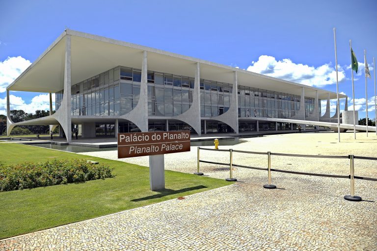 Prédio branco com uma rampa na frente, um espelho d'água ao redor, um gramado à esquerda, uma ampla calçada de pedras brancas à frente, bandeiras hasteadas e uma placa escrito: Palácio do Planalto 