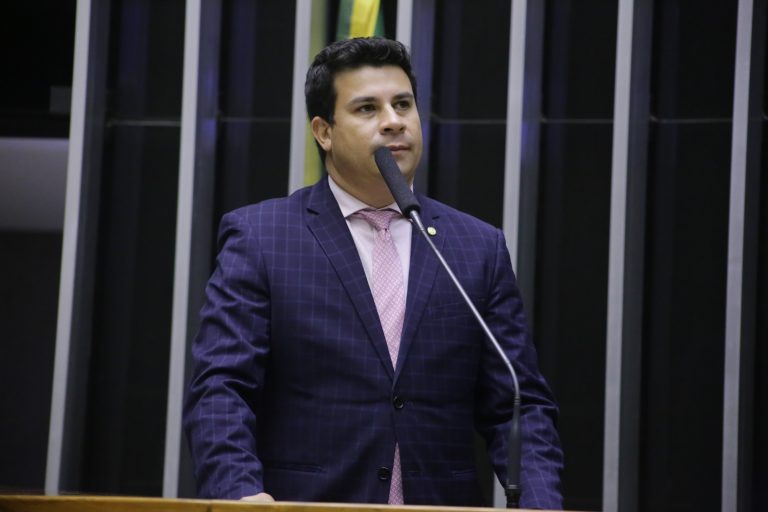 Deputado Carlos Veras fala ao microfone. Ele é branco, tem o cabelo escuro, usa um terno azul-marinho e camisa e gravata rosas