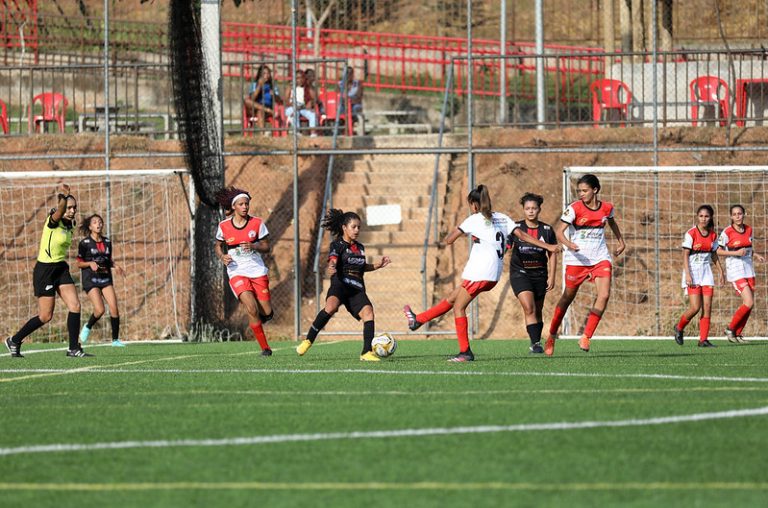 Mulheres jogam futebol num campo de grama num dia de sol. Um time está com short vermelho e camisa branca e o outro está com uniforme preto