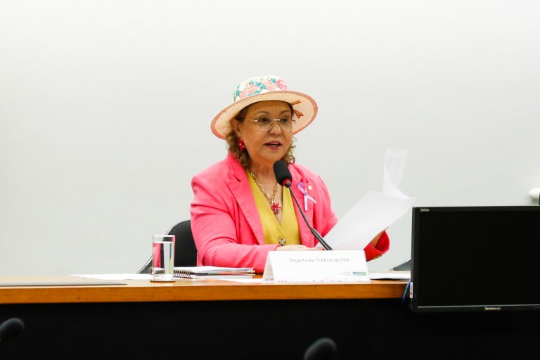Deputada Tereza Nelma fala ao microfone. Ela é branca, usa chapéu e um blazer rosa