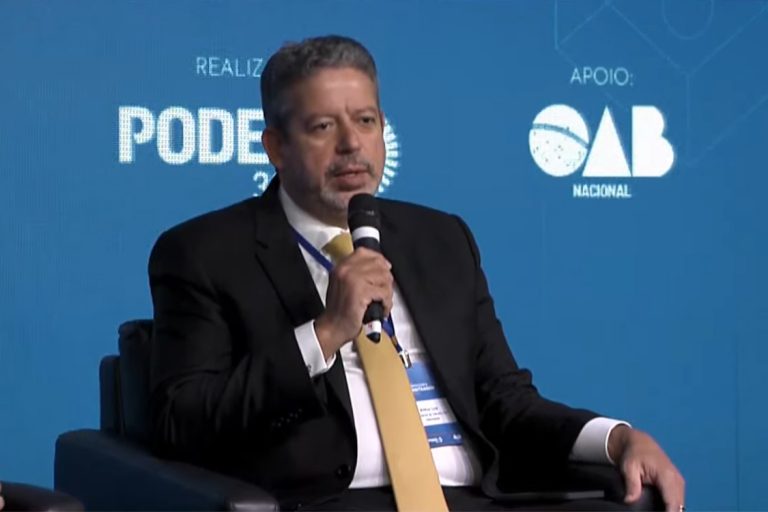 Deputado Arthur Lira é um homem branco com cabelo escuro, barba e bigode, usa um terno escuro, segura um microfone e está sentado à frente de um fundo azul