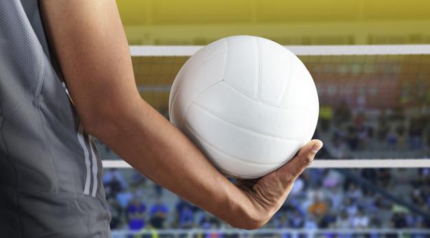 Foto cortada de um homem segurando uma bola de vôlei branca, ao fundo há uma rede de vôlei e o público desfocados