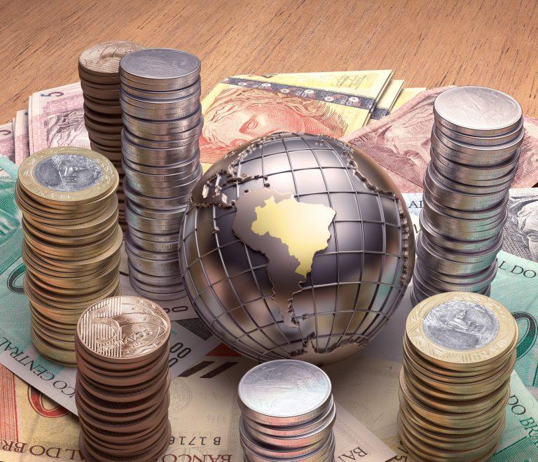 Globo terrestre com o Brasil em dourado em cima de notas de dinheiro e ao lado de pilhas de moedas