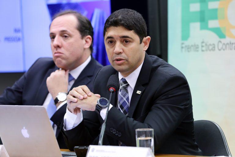 Dois homens estão sentados de terno e um deles fala ao microfone