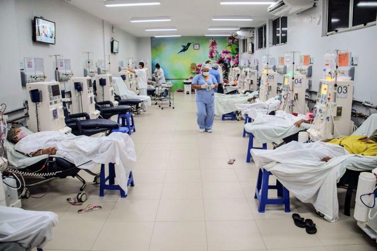 Sala com vários pacientes deitados fazendo hemodiálise e uma enfermeira caminhando entre eles