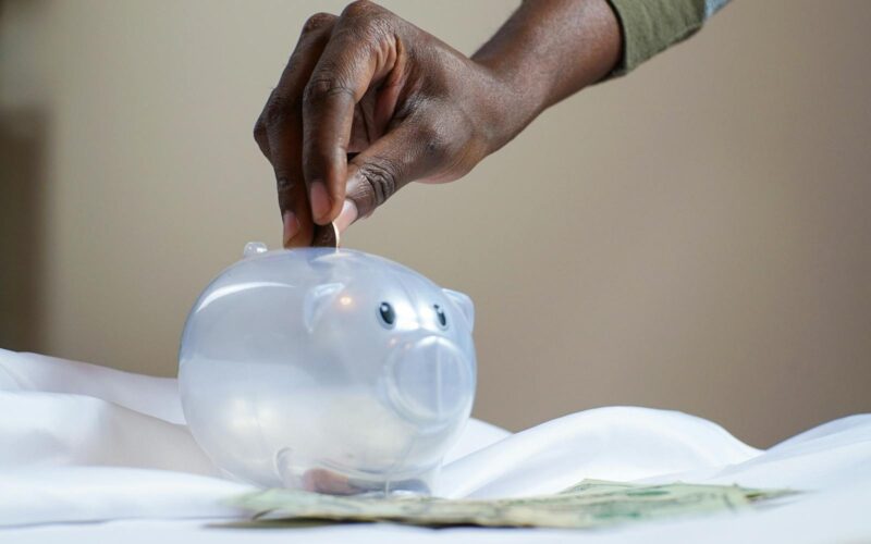 Close-up of a person's hand placing coins into a transparent piggy bank to save money.